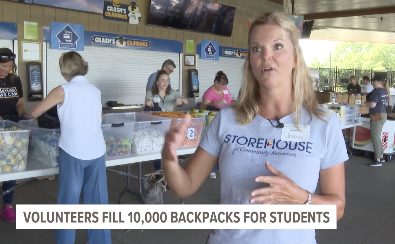 A blonde woman wearing a gray shirt explains how volunteers are filling backpacks for students. 
